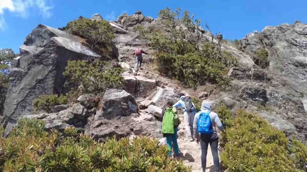 Hikers climbing the rocky summit trail of Volcán Barú in Panama under a clear blue sky, showing the steep volcanic terrain and lush vegetation.